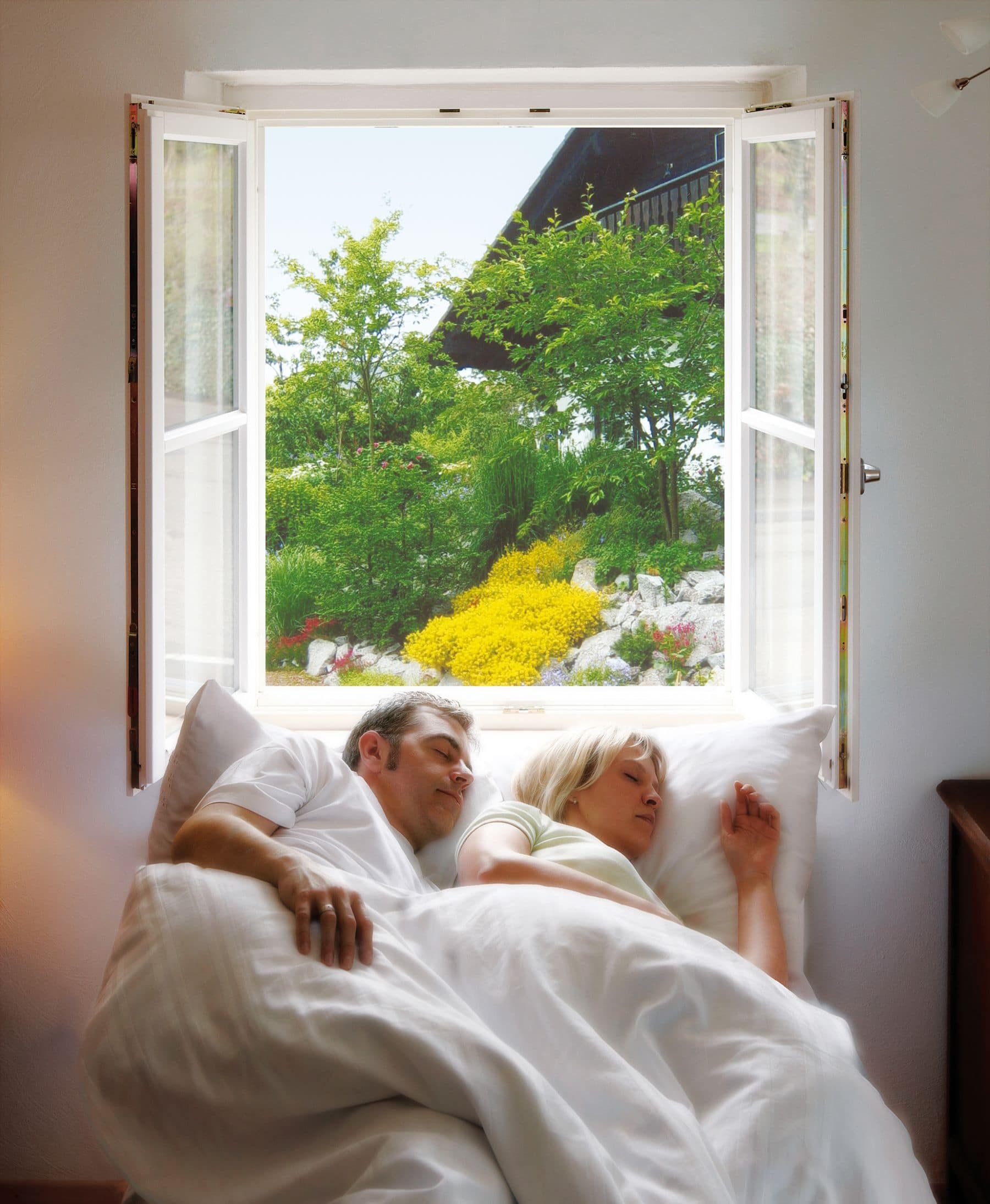 Ein Paar ruht sich im Bett aus. Das offene, durch Insektenschutz geschützte Fenster bietet einen Blick auf einen Garten und die Außenansicht eines Gebäudes.