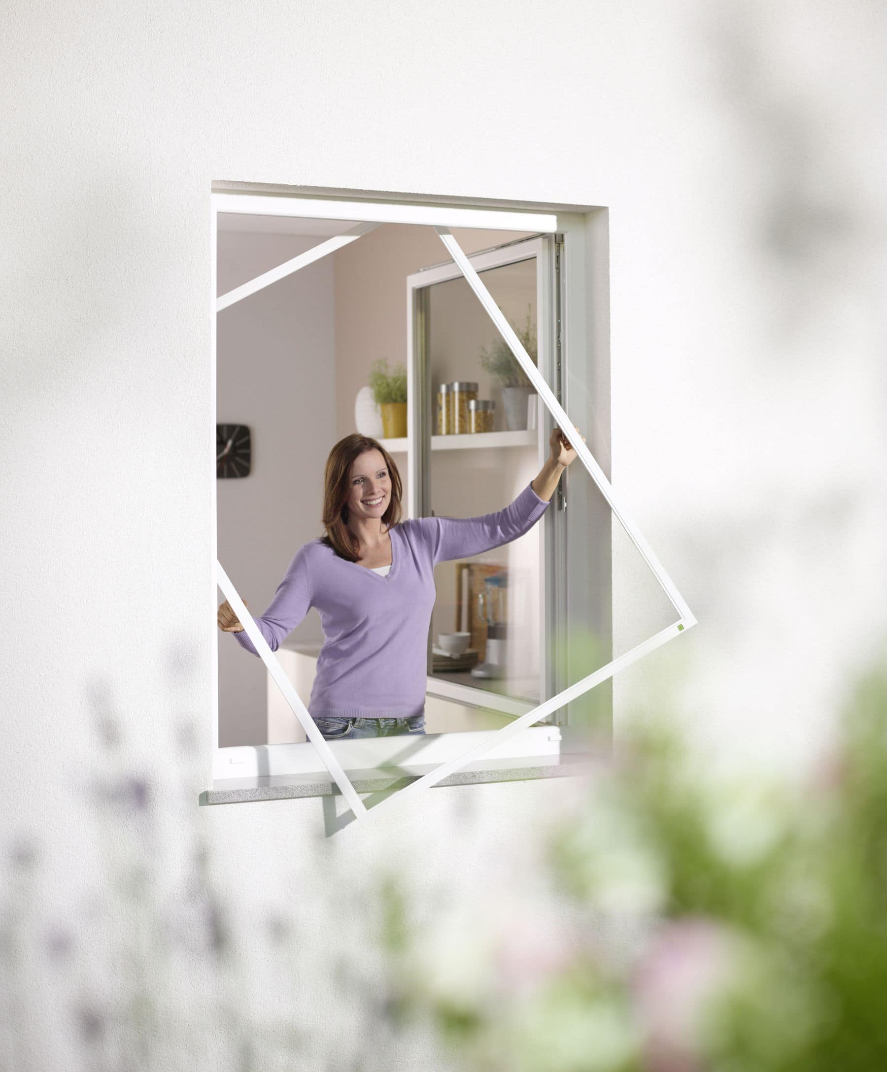 Eine lächelnde Frau installiert von der Innenseite eines Raumes aus einen Insektenschutz an einem offenen Fenster.