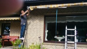 Mann installiert eine Markise vor einem Fenster und steht auf einem Hocker mit einer Leiter in der Nähe.