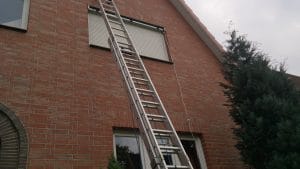 Eine ausziehbare Leiter lehnt an einem Backsteinhaus neben einem geschlossenen Fenster mit Metallläden.