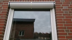 Außenansicht eines geschlossenen doppelt verglasten Fensters mit Rollladen an einer Ziegelwand, in dem sich ein bewölkter Himmel und ein Teil des Nachbarhauses spiegeln.